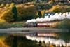 A steam train passing between a house and a lake with autumn trees behind.