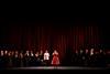 A cast of performers on stage in front of a red curtain.