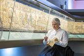 Woman doing embroidery while sat next to Britain's Bayeux Tapestry