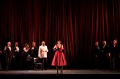 A cast of performers on stage in front of a red curtain.
