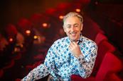 Alan Cumming sat in red theatre seat smiling at camera
