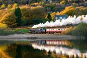A steam train passing between a house and a lake with autumn trees behind.