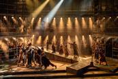 Cast members on stage performing around scaffolding building