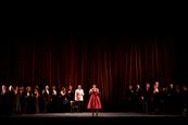 A cast of performers on stage in front of a red curtain.