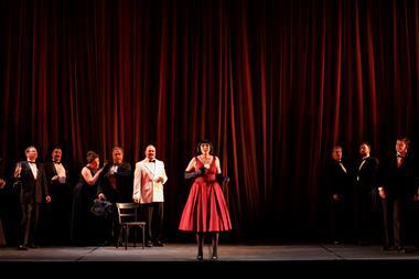 A cast of performers on stage in front of a red curtain.