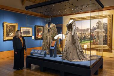 A woman stands beside a display case containing Coronation outfits