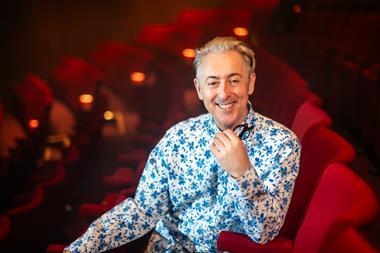 Alan Cumming sat in red theatre seat smiling at camera
