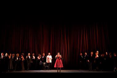 A cast of performers on stage in front of a red curtain.