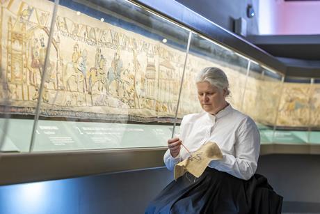 Woman doing embroidery while sat next to Britain's Bayeux Tapestry