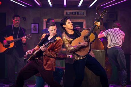 Two male cast members playing acoustic guitars on stage in front of a piano.