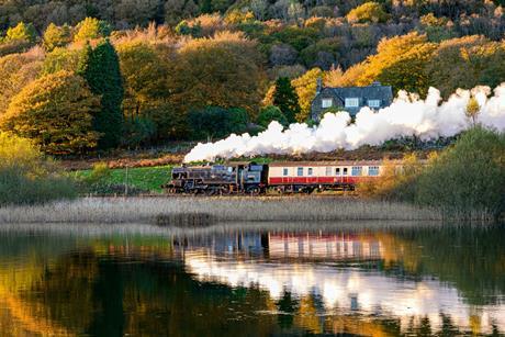 A steam train passing between a house and a lake with autumn trees behind.