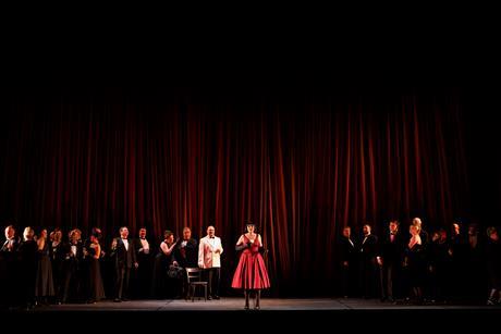 A cast of performers on stage in front of a red curtain.