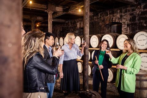 A group of young people on a tour of the Bushmills Whiskey Distillery. Each of them has a glass of whiskey and they're raising their glasses and smiling at each other.