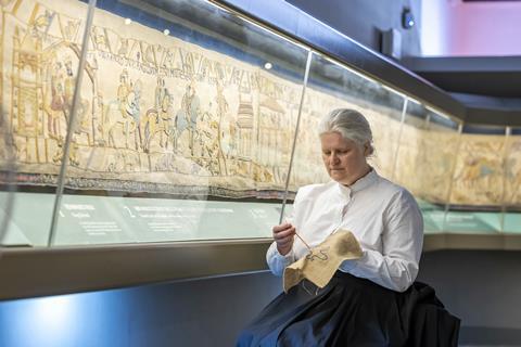 Woman doing embroidery while sat next to Britain's Bayeux Tapestry