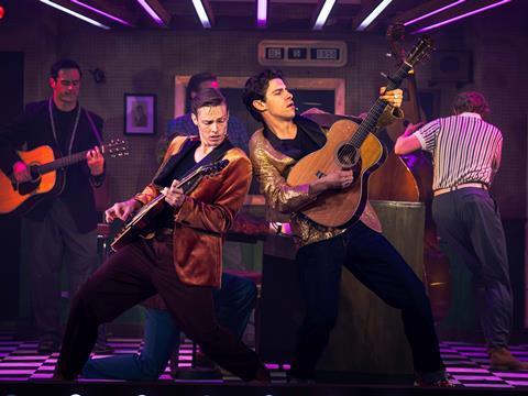 Two male cast members playing acoustic guitars on stage in front of a piano.